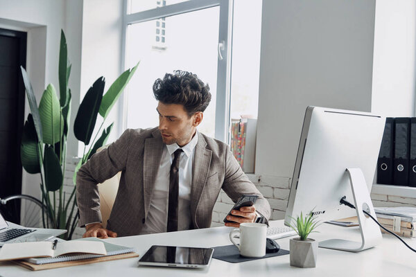 Thoughtful young man holding smart phone while sitting at his working place in office
