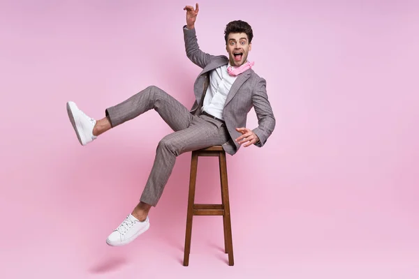 Joyful young man in full suit having fun while sitting on the chair against pink background