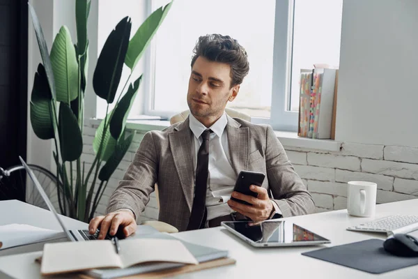 Confident Young Man Using Laptop Holding Smart Phone While Sitting — Photo