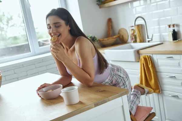 Beautiful Young Woman Eating Cookies While Sitting Kitchen Counter — Stockfoto
