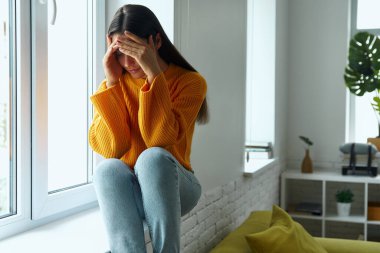 Depressed young woman covering face with hands while sitting on the window sill at home