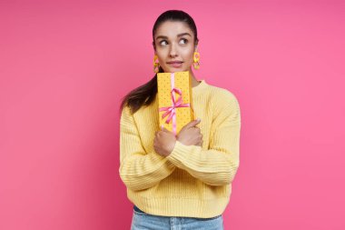 Dreamful young woman holding gift box and looking away against colored background
