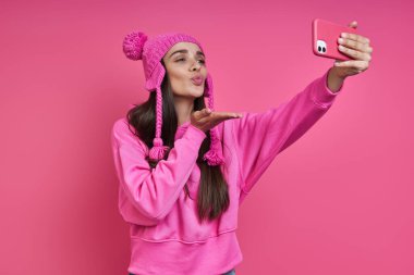 Playful young woman in funky hat making selfie and blowing a kiss against pink background