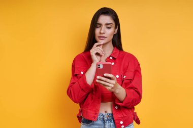 Thoughtful young woman using smart phone while standing against yellow background