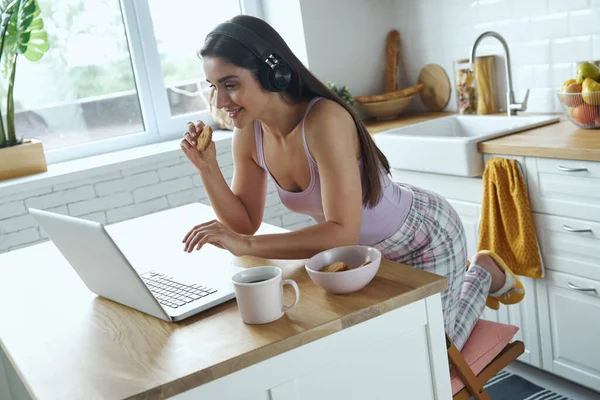 Attractive Woman Headphones Using Laptop Eating Cookies While Sitting Kitchen — Stockfoto