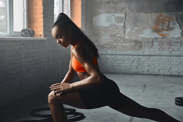 Beautiful young African woman in sports clothing warming up before training in gym