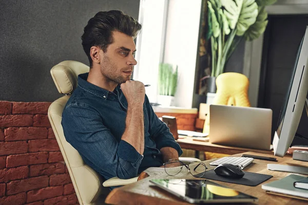 Concentrated Young Man Holding Hand Chin While Sitting His Working — Stockfoto