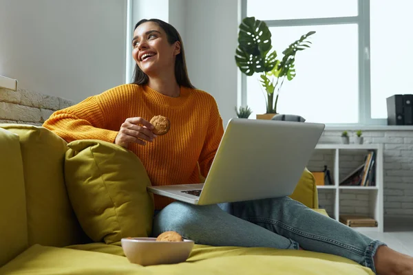 Relaxed Young Woman Using Laptop Enjoying Cookies While Sitting Couch — Stockfoto