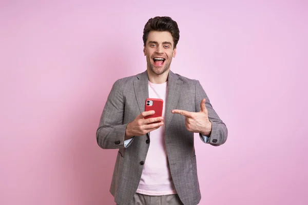 Excited young man in suit pointing smart phone while standing against pink background