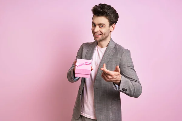 Happy young man in suit pointing gift box while standing against pink background