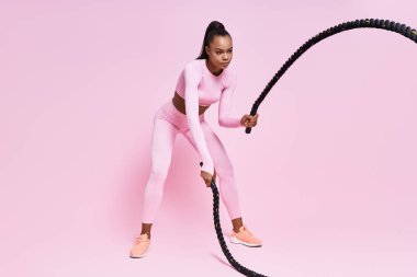 Concentrated young African woman exercising with battle ropes against pink background