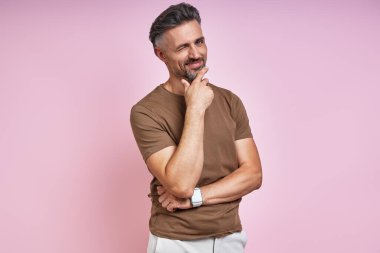 Handsome mature man holding hand on chin and winking while standing against pink background