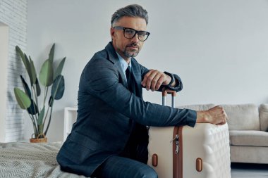 Confident mature man in formalwear leaning on his luggage and looking at camera while sitting on bed