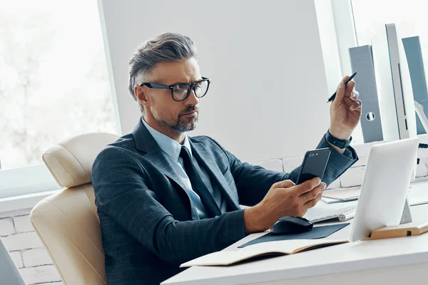 Confident Businessman Holding Smart Phone Pointing Computer Monitor While Sitting — Stock fotografie