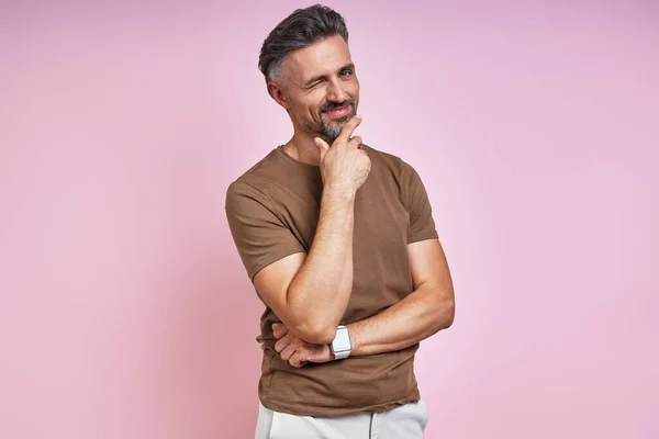 Handsome mature man holding hand on chin and winking while standing against pink background