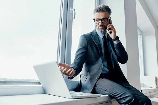 Confident Businessman Talking Mobile Phone Using Laptop While Sitting Window — Stock fotografie