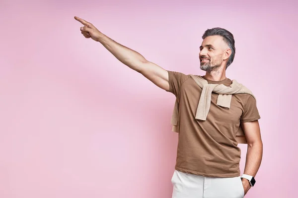 Handsome mature man pointing away and smiling while standing against pink background