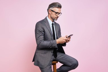 Confident mature man in formalwear using smart phone while sitting against pink background