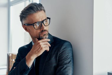 Thoughtful mature man in formalwear holding hand on chin while looking through a window