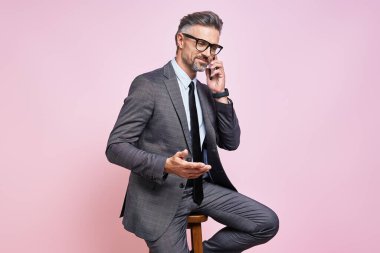 Handsome mature man in formalwear talking on mobile phone and gesturing against pink background