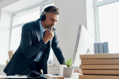 Thoughtful businessman in headphones looking at the computer while standing in office