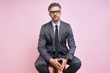 Handsome mature man in formalwear keeping hands clasped while sitting against pink background