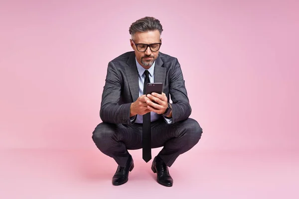 Confident mature man in full suit looking at camera while sitting against pink background