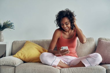 Evdeki kanepede dinlenirken akıllı telefon kullanan güzel bir Afrikalı kadın.