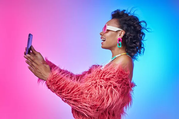 Şık ceketli, akıllı bir telefon tutan ve renkli arka planda gülümseyen Afrikalı bir kadın.