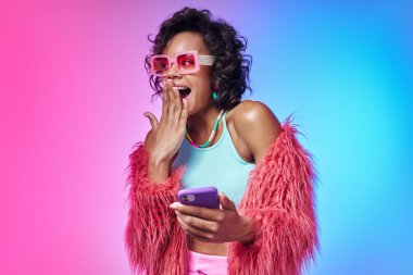 Moda kıyafetleri içinde heyecanlı Afrikalı kadın akıllı telefonunu gösteriyor ve renkli arka planda gülümsüyor.