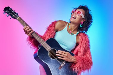 Kabarık ceketli, gitar çalan ve renkli arka planda gülümseyen güzel Afrikalı kadın.