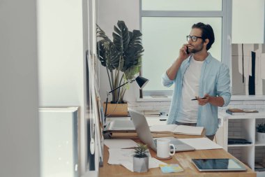 Ofisteki iş yerinin yanında duran kendine güvenen genç adam cep telefonuyla konuşuyor.