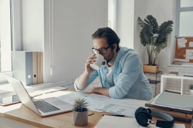 Yakışıklı, kahve içen ve ofiste otururken dizüstü bilgisayara bakan genç adam.