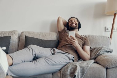 Kulaklıklı neşeli genç adam evde kanepede dinlenirken elinde akıllı bir telefon tutuyor.