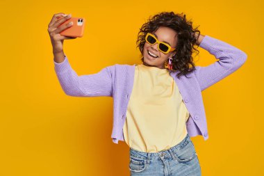 Ürkütücü gözlüklü mutlu Afrikalı kadın sarı arka planda selfie çekiyor.