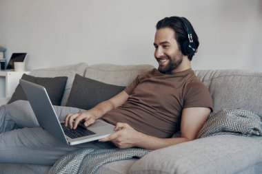 Kulaklıklı yakışıklı genç adam evdeki kanepede dinlenirken akıllı telefon kullanıyor.