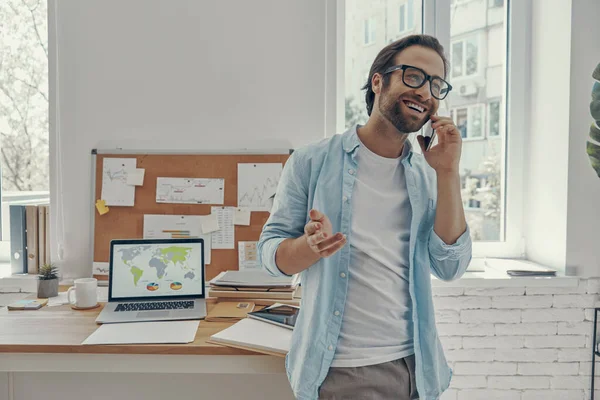 Confident young man talking on mobile phone while standing near his working place in office