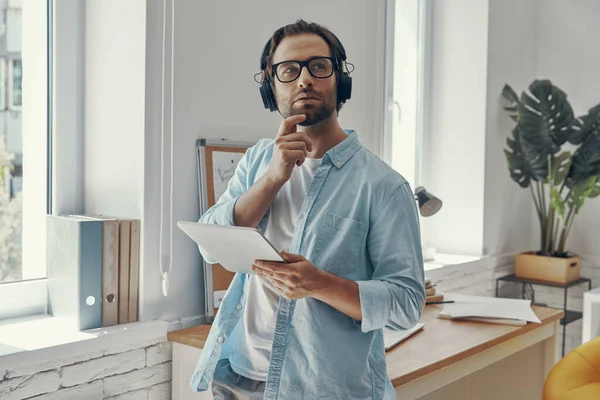 オフィスに立っている間にデジタルタブレットを保持してヘッドフォンで思慮深い若い男 — ストック写真
