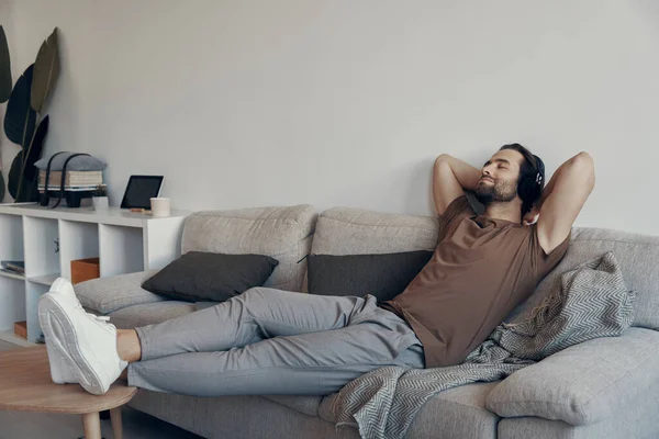 Hezký Mladý Muž Sluchátkách Drží Ruce Hlavou Zatímco Relaxuje Gauči — Stock fotografie