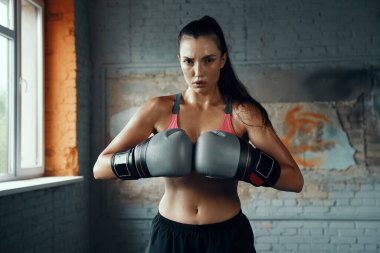 Boks eldivenli, kendine güvenen genç bir kadın spor salonunda kameraya bakıyor.