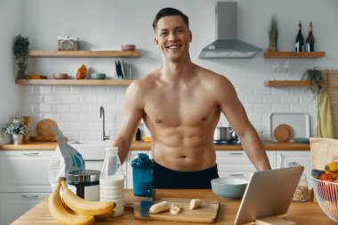 Yakışıklı genç adam kameraya bakıyor ve mutfakta sağlıklı yemek hazırlarken gülümsüyor.