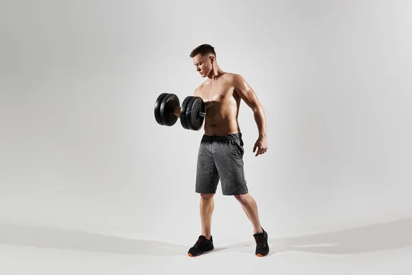Concentrated young man with perfect body exercising with dumbbell against white background