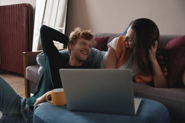 Mutlu genç çift evde dinlenirken dizüstü bilgisayar kullanıyor.