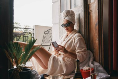 Bornozlu güzel bir kadın balkonda otururken akıllı telefon kullanıyor.