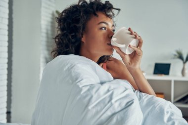 Güzel Afrikalı kadın yatakta tembelce vakit geçirirken sabah kahvesinin tadını çıkarıyor.