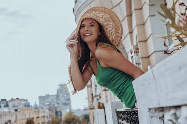 Güzel genç bayan zarif şapkasını düzeltiyor ve balkonda dikilirken manzaraya bakıyor.
