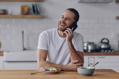 Mutfakta yemek yerken cep telefonuyla konuşan mutlu genç adam.