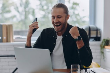İşyerinde otururken dizüstü bilgisayara bakıp el kol hareketi yapan mutlu genç adam.