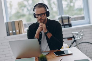 Kulaklıklı konsantre bir adam ofisteki iş yerinde laptopuna bakıyor.