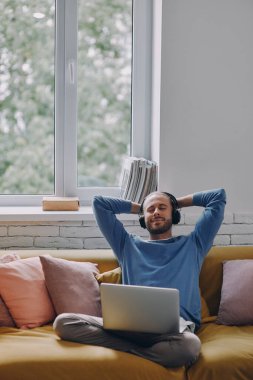 Evde kanepede otururken kulaklıklı, el ele tutuşan rahat genç bir adam.